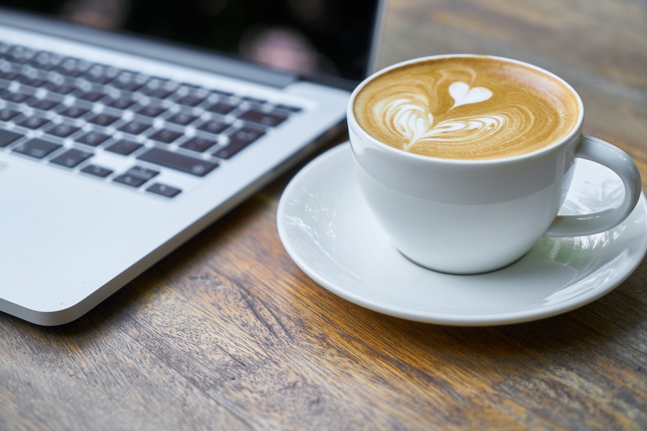 a frothy cup of coffee on a table beside a laptop