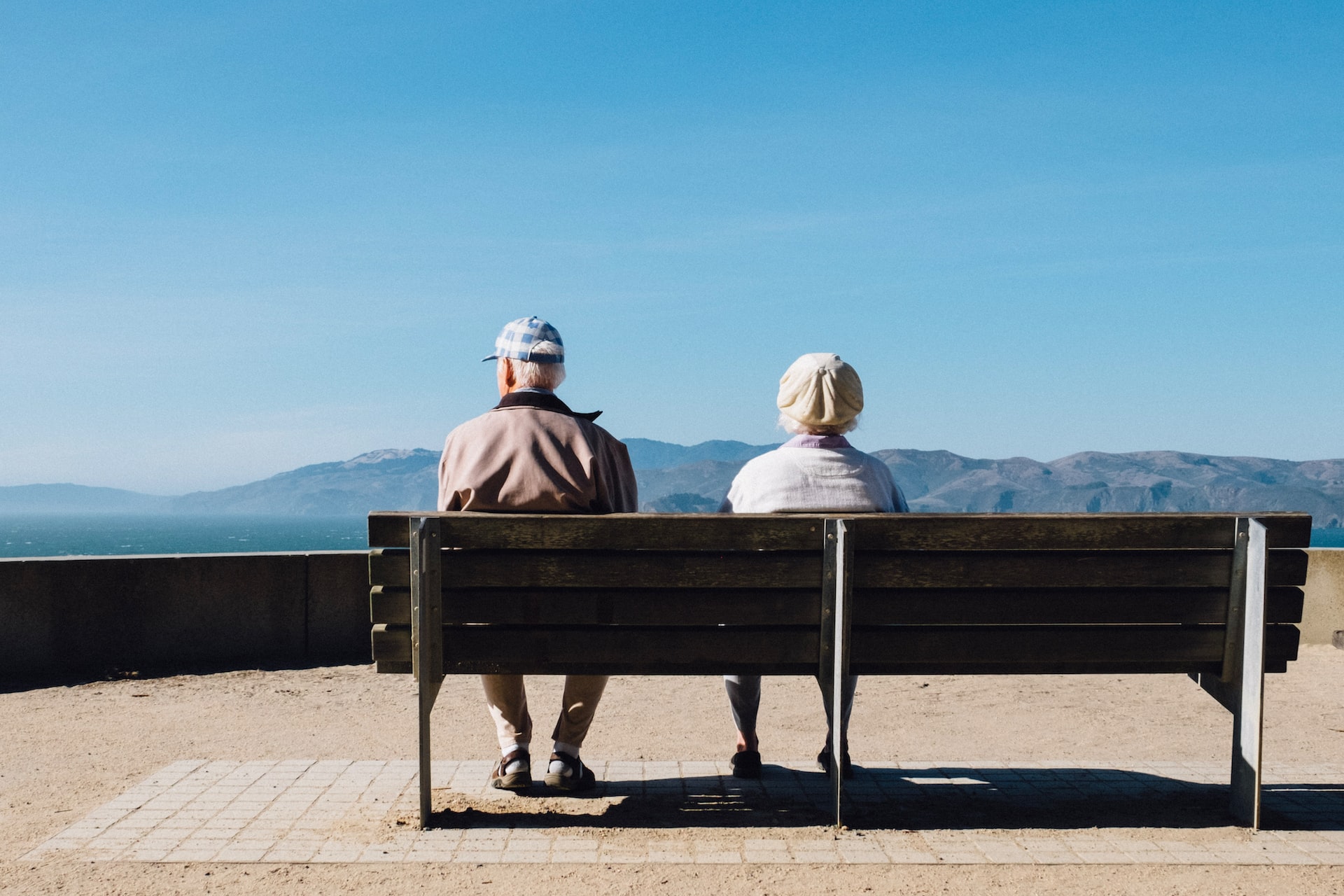 two elderly people sitting on a bench by the water in front of mountains